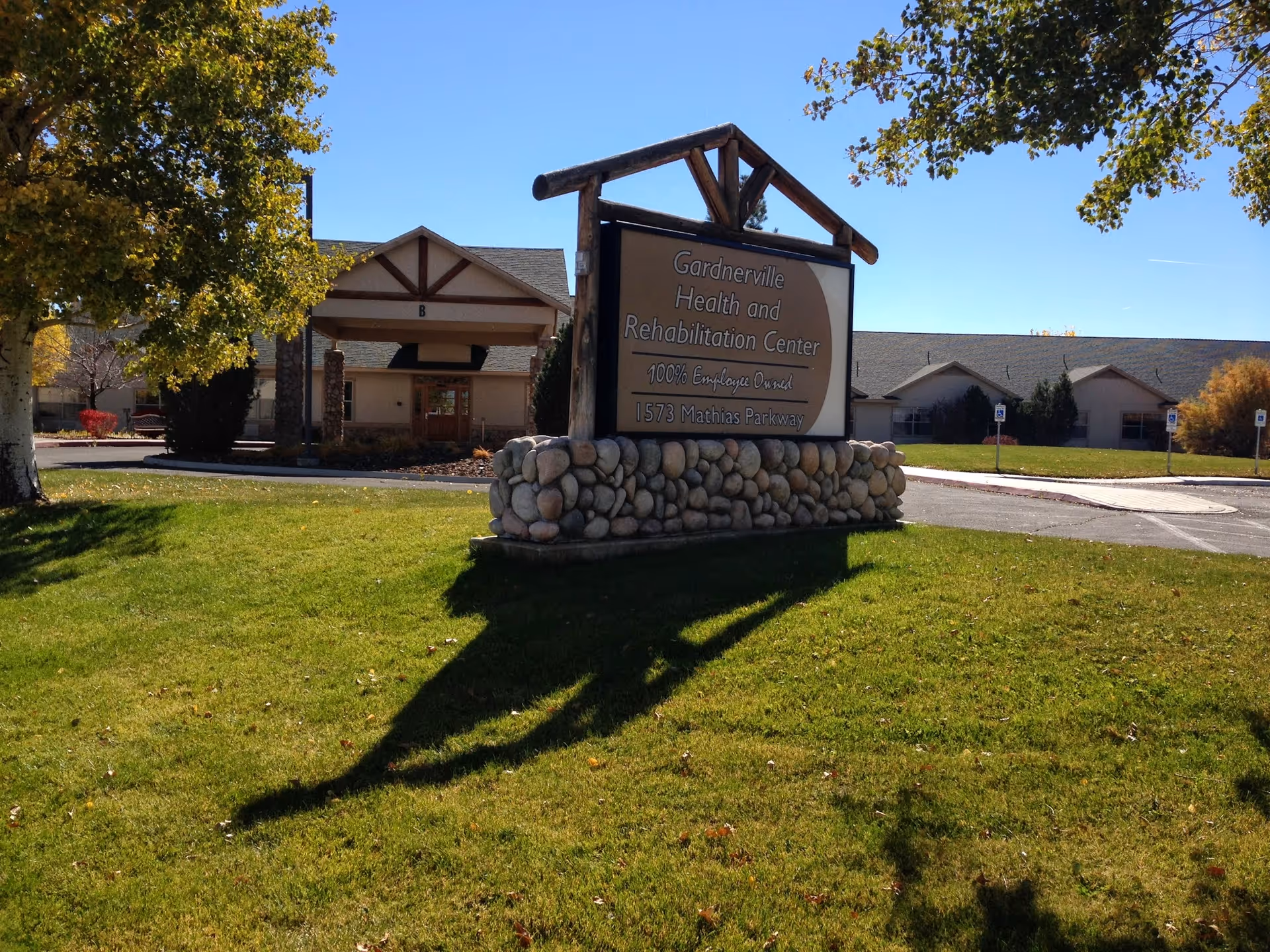 Outdoor view of Gardnerville Health and Rehabilitation Center showing a large sign with the facility name and address on a stone base, with the building and entrance visible in the background under a clear blue sky and surrounded by green grass and trees.
