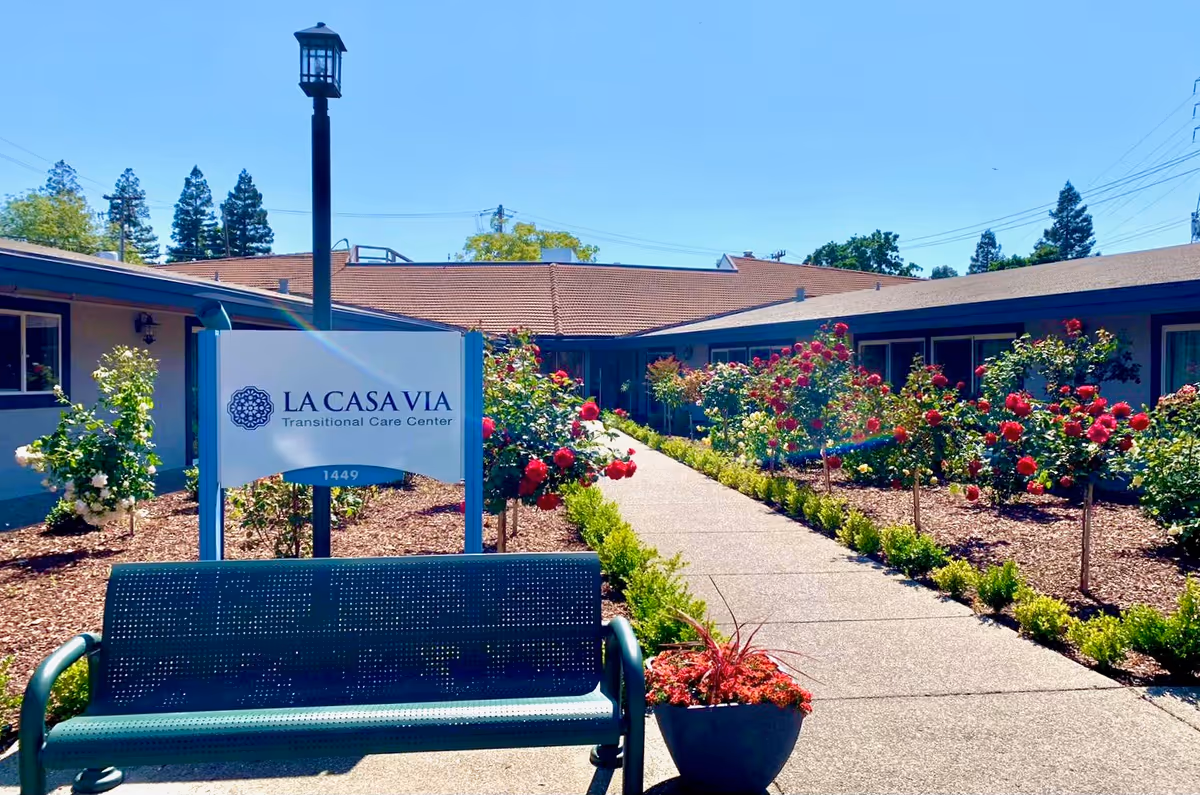 Outdoor view of La Casa Via Transitional Care Center featuring a pathway lined with red and white rose bushes, a green bench in front of a sign with the facility's name, and a building with a brown roof in the background under a clear blue sky.