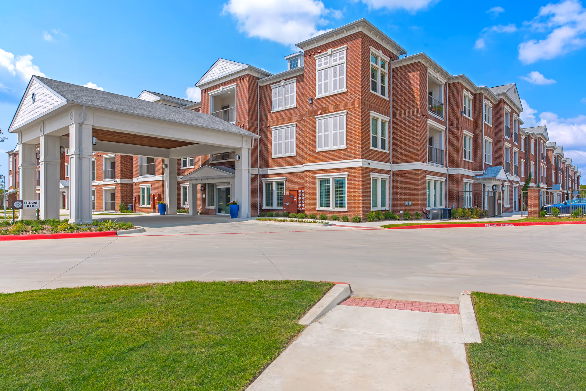 Exterior view of a multi-story brick building with white trim and multiple windows under a blue sky with some clouds. The building has a covered entrance with large white columns and a sign indicating the leasing office. There is a paved driveway and well-maintained green grass in the foreground.