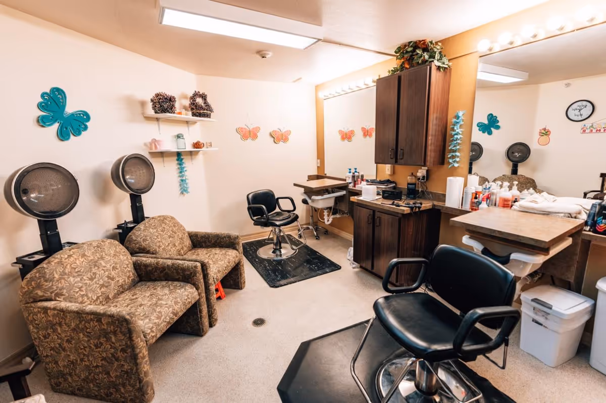 Interior of a hair salon room in an assisted living facility with two patterned armchairs under hair dryers, two black salon chairs in front of a large mirror, wooden cabinets, and butterfly wall decorations.