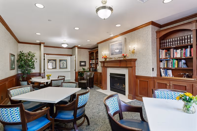 Bright senior living common room with several dining tables and chairs, a fireplace, built-in bookshelves, and a small seating area.