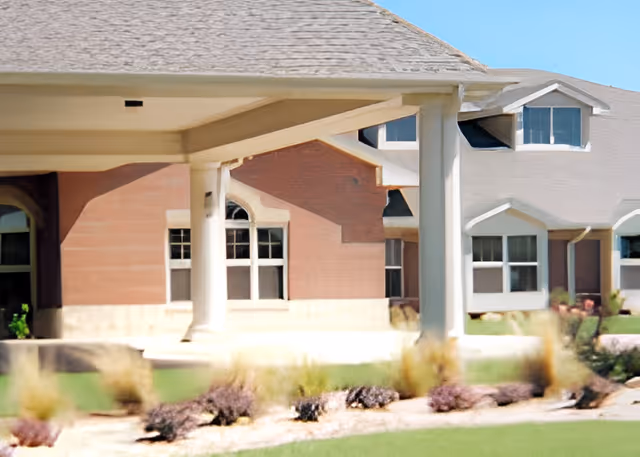 Covered front entrance with columns, windows, and landscaped lawn in front of a multi-windowed brick-and-siding building.