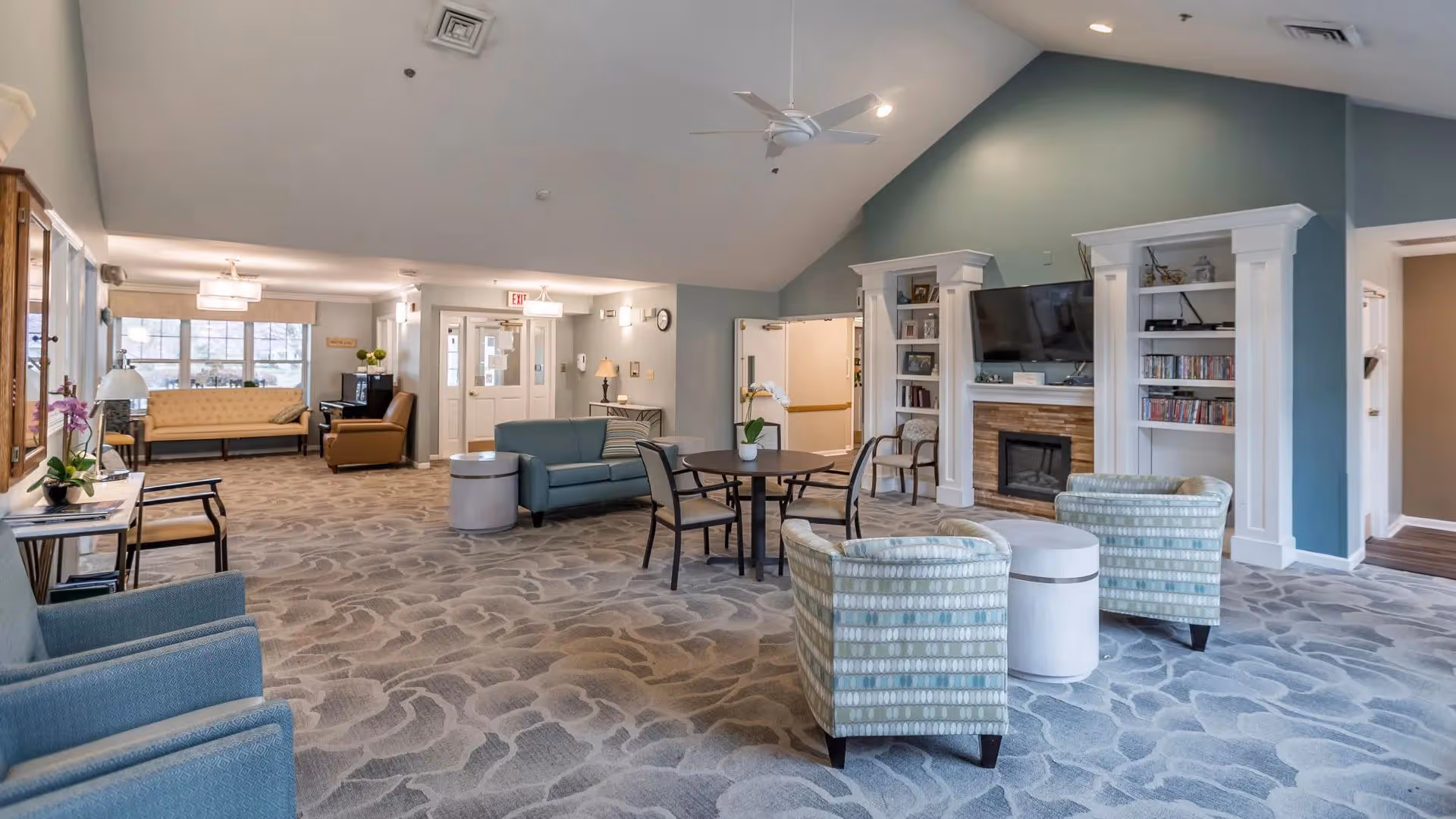 A spacious senior living facility common area with patterned carpet, multiple seating options including armchairs, sofas, and a round table with chairs. There is a fireplace with a mounted TV above it, built-in shelves with books and decor, and large windows letting in natural light. The walls are painted in soft colors, and the ceiling is vaulted with a ceiling fan.