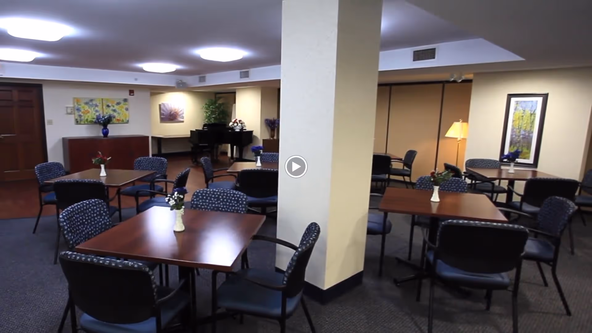 Community dining room with several square wooden tables and upholstered chairs arranged around a central column, small vases with flowers on the tables, and a piano in the background.