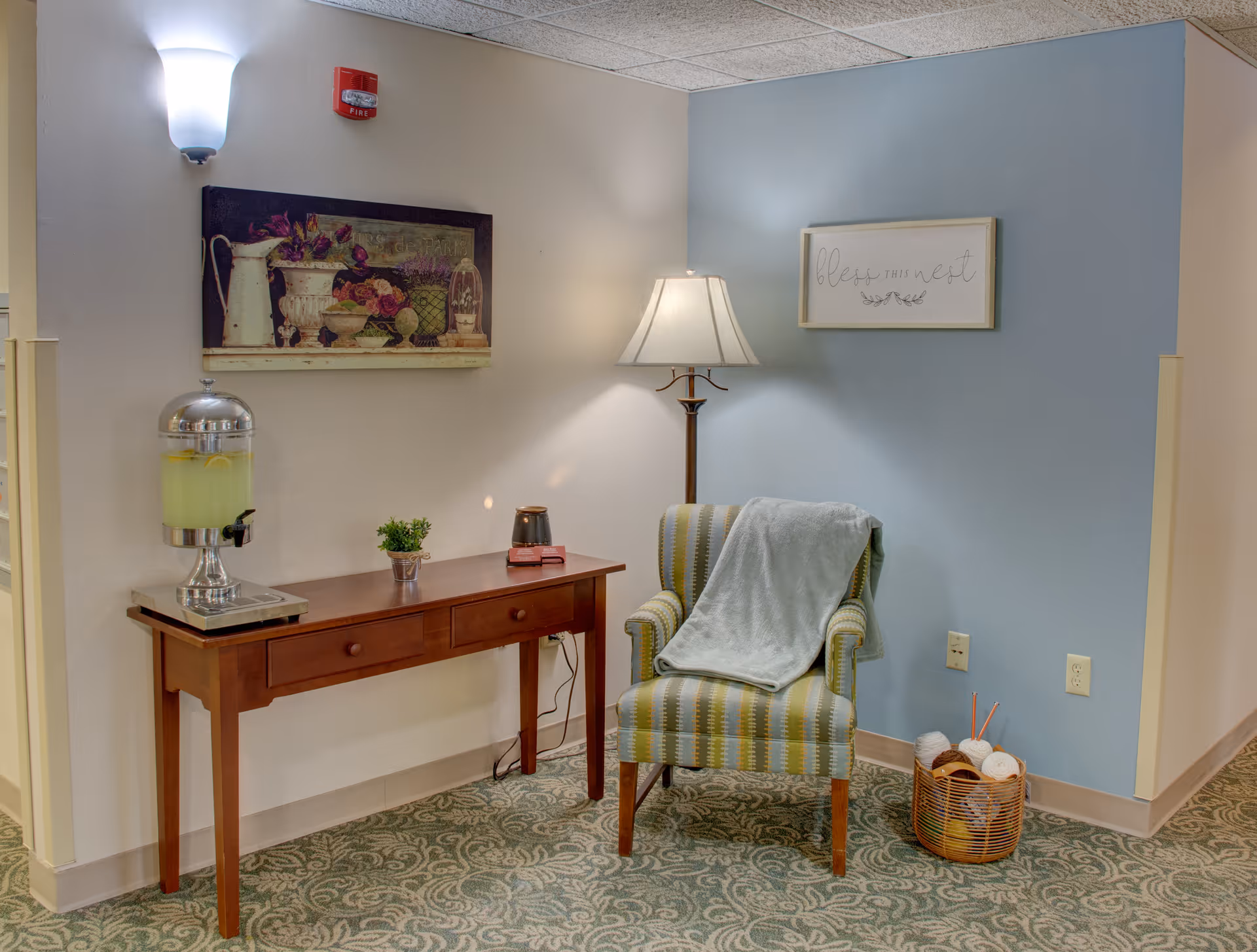 A cozy corner in a senior living facility with a striped armchair draped with a light blue blanket, a floor lamp beside it, and a basket filled with yarn and knitting needles on the floor. A wooden console table holds a beverage dispenser with lemonade, a small potted plant, and a candle. The walls are decorated with two framed artworks, one with flowers and the other with the words 'bless our nest'.