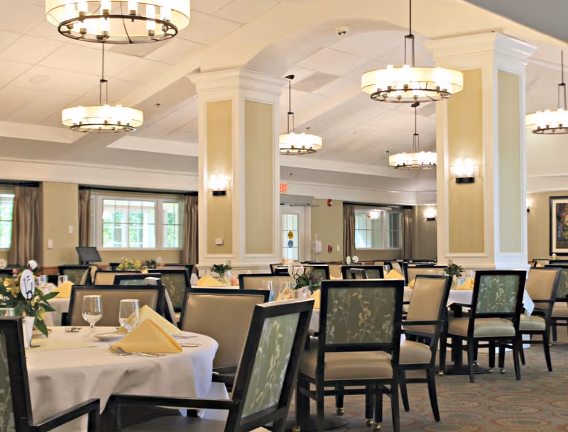 A spacious dining room with multiple round tables covered with white tablecloths, each set with folded yellow napkins, glassware, and silverware. The chairs have dark wooden frames with light-colored floral upholstery. The room features large windows with curtains, beige walls with white trim, and several modern circular chandeliers hanging from the ceiling providing warm lighting.
