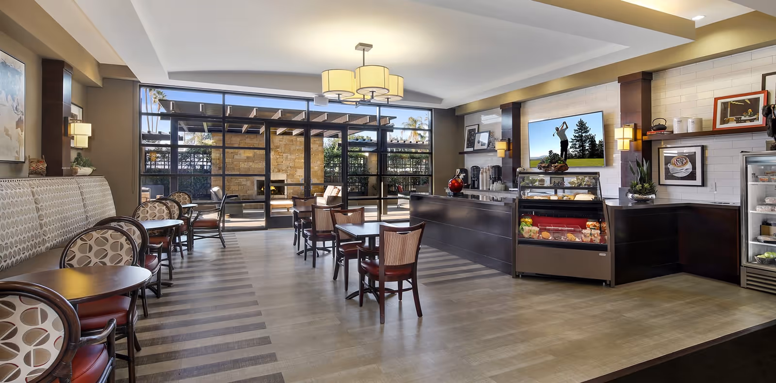 A bright and modern dining area with several tables and chairs arranged neatly. There is a counter with a display case containing food items, and a large flat-screen TV mounted on the wall showing a golfer. Large glass doors lead to an outdoor patio area with seating and a stone fireplace. The room features warm lighting and tasteful decor.