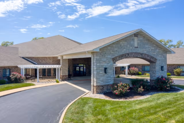 Exterior view of Cedarhurst of Breese facility showing a large covered entrance with stone and brick facade, surrounded by well-maintained green lawns and flowering bushes under a partly cloudy blue sky.