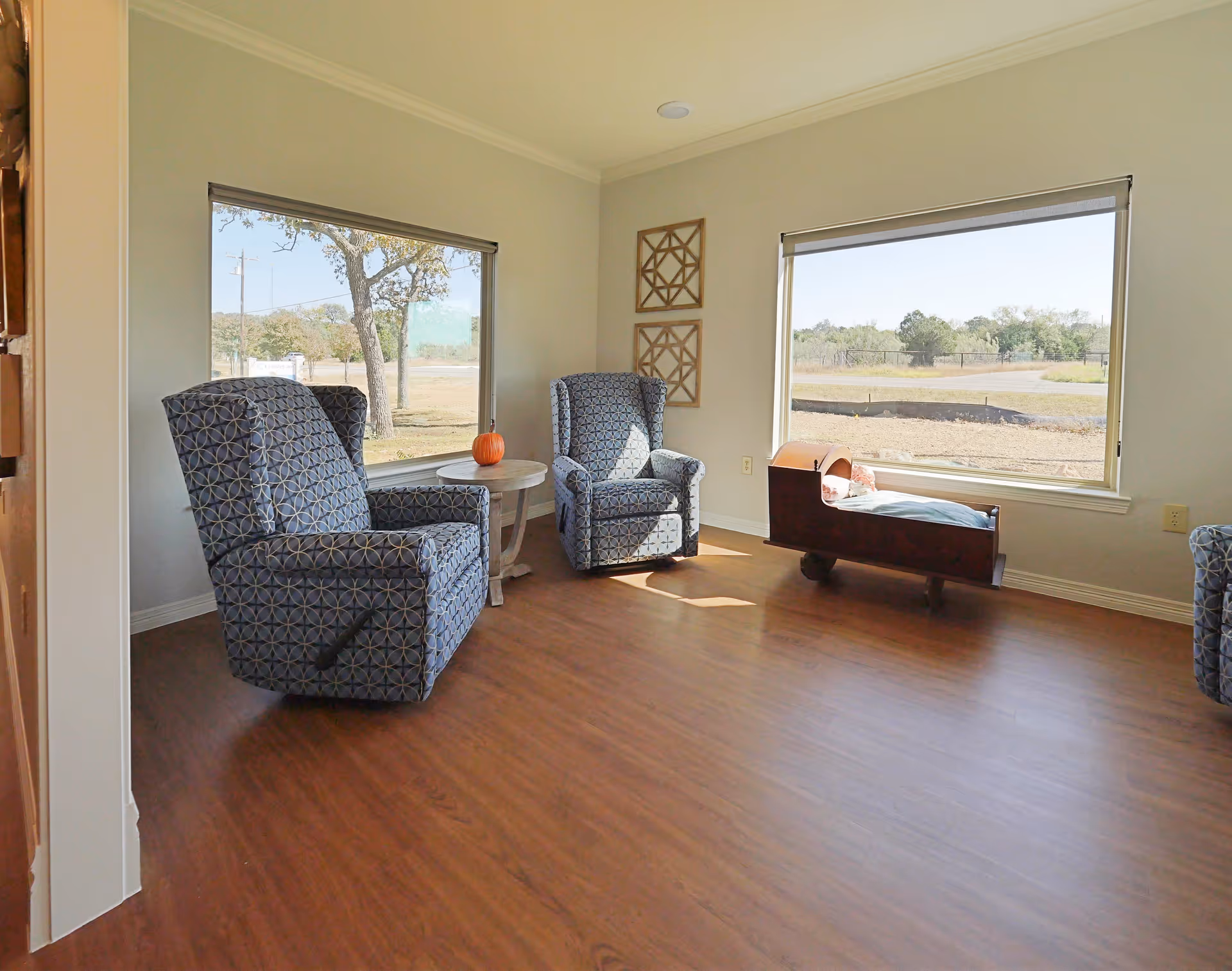 A bright room with two patterned armchairs and a small round table between them, placed near large windows showing an outdoor view with trees and open land. There is a wooden cradle with a pillow near one window and two decorative wooden wall hangings on the wall.