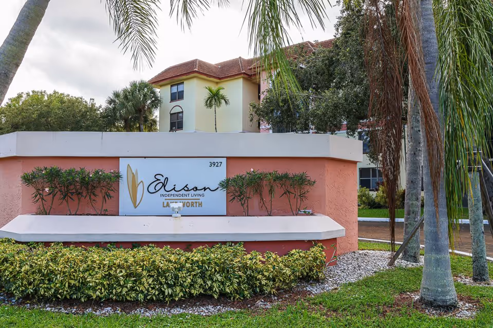 Outdoor view of the entrance sign for Elison Independent Living of Lake Worth, surrounded by palm trees and greenery, with a multi-story building in the background.
