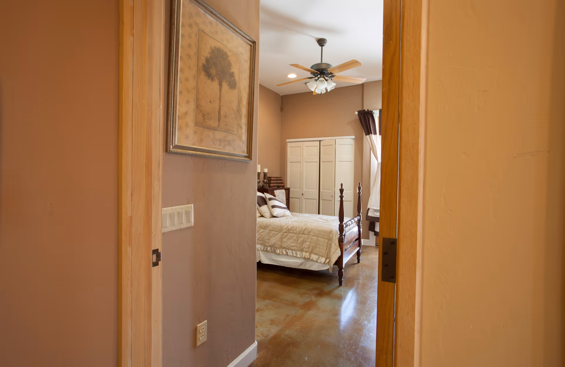 View through an open doorway into a bedroom with a wooden bed frame, beige bedding, a ceiling fan with lights, a window with curtains, and a white wardrobe. The walls are painted in warm tones and there is a framed picture hanging on the wall outside the bedroom.