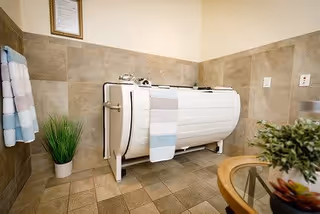 A spacious bathroom with beige tiled walls and floor featuring a white medical-style bathtub with safety rails. Towels hang on the wall, and there are two potted plants adding greenery to the room. A glass table with a plant is partially visible in the foreground.