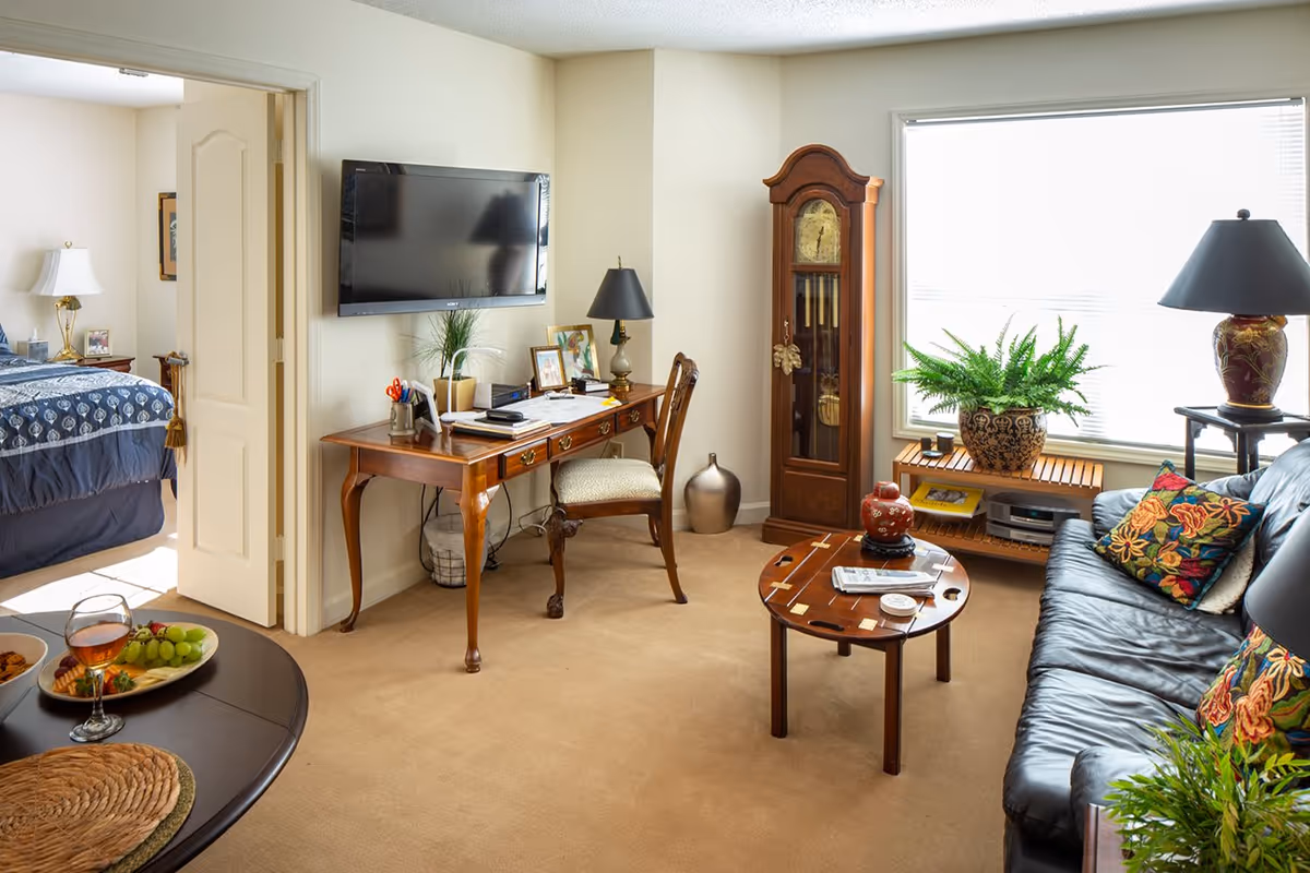 A cozy living room with a black leather couch adorned with colorful floral pillows, a round wooden coffee table with decorative items, a wooden desk with a chair and table lamp, a wall-mounted flat-screen TV, and a tall grandfather clock near a large window with blinds. The room has beige carpet and walls. A bedroom with a bed and nightstand is visible through an open door.