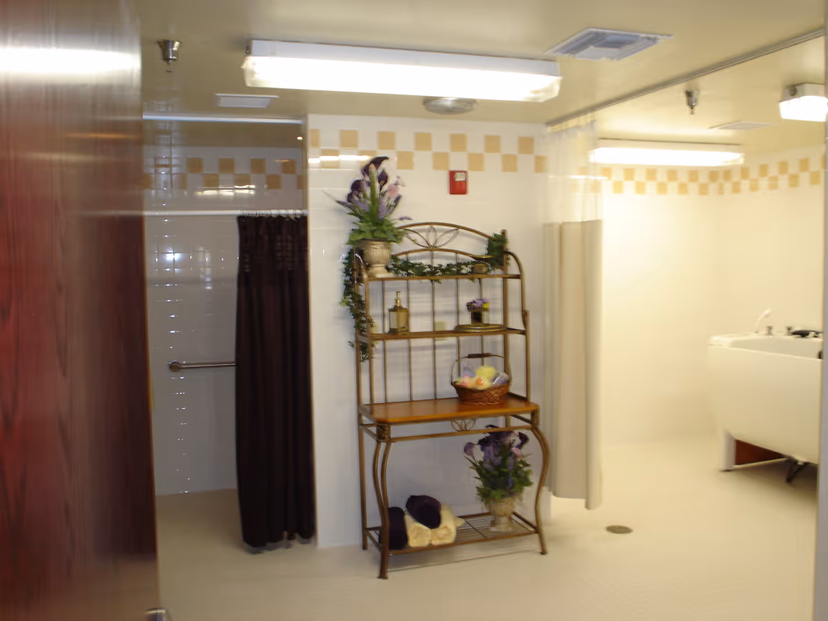 Interior view of a bathroom area with white tiled walls featuring a yellow checkered border near the ceiling. There is a metal shelving unit decorated with plants, towels, and a basket. A dark shower curtain is partially visible on the left side, and a white bathtub with a curtain is on the right side. The room is well-lit with fluorescent ceiling lights.