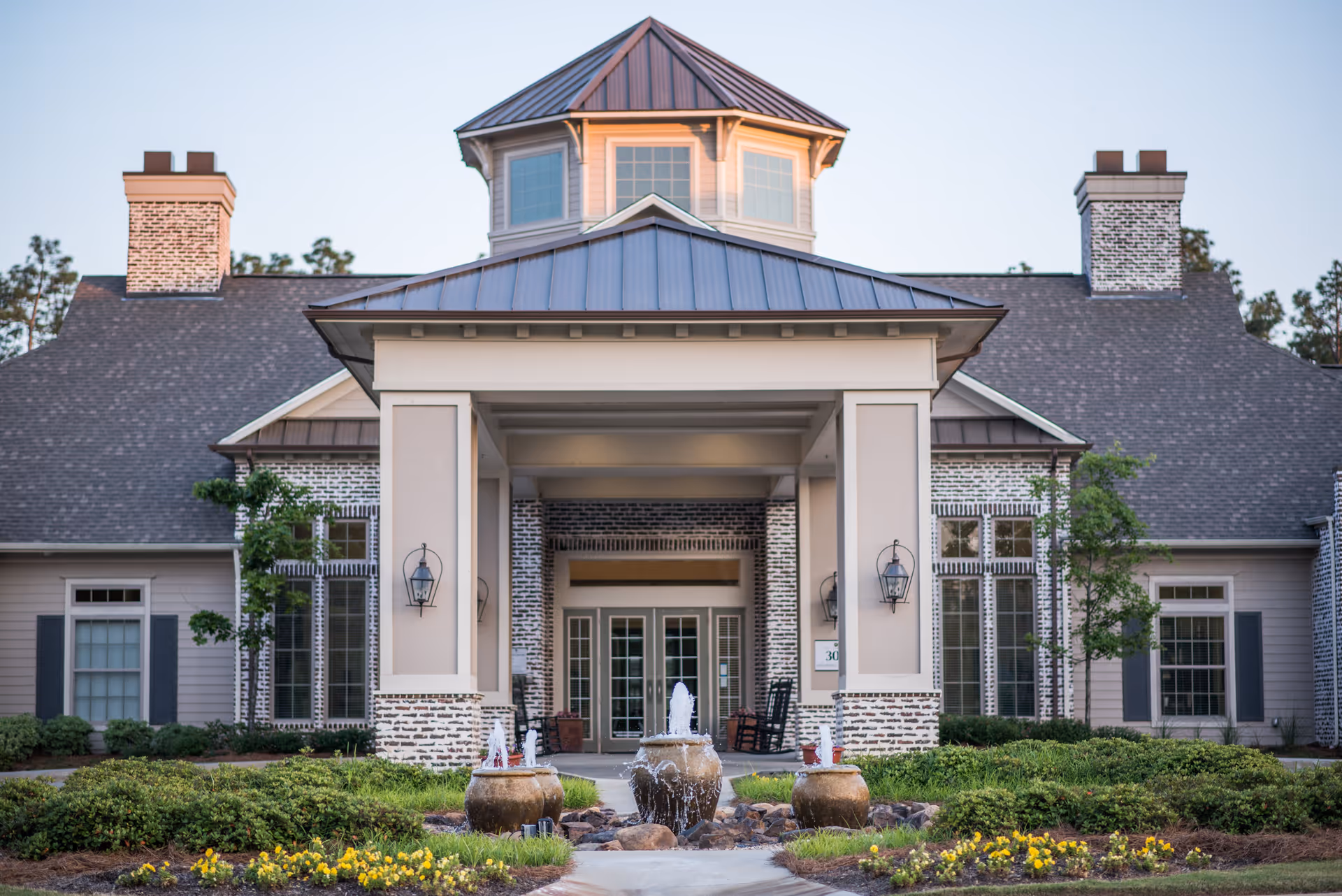 Front exterior view of The Claiborne at Hattiesburg Independent Living building with a covered entrance, three water fountains, landscaped bushes, and flowers in the foreground.