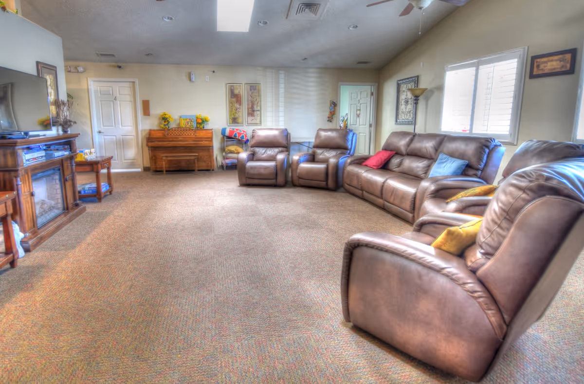 A spacious living room with brown leather recliners and a matching sofa arranged around the room. There is a wooden TV stand with a flat-screen TV on the left side, a wooden piano against the back wall, and a small table with a lamp near the window on the right. The room has beige walls, carpeted floor, and windows with white shutters letting in natural light.