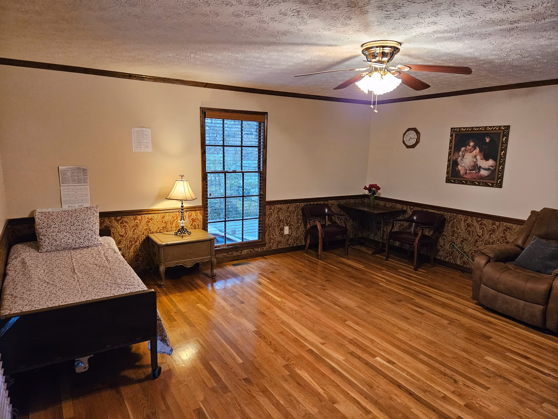 Spacious senior facility bedroom with a single bed and nightstand, a seating area with chairs and a recliner, hardwood floors and a ceiling fan.