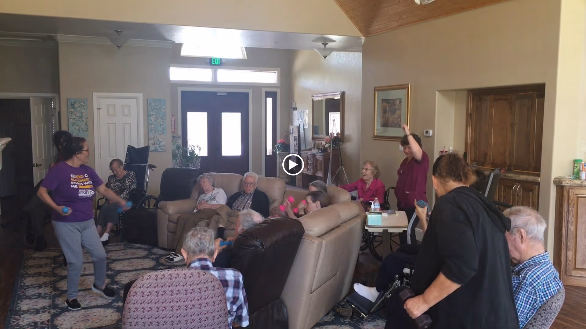 A group of elderly people seated in a living room area participating in a group activity led by two caregivers. The room has comfortable seating including sofas and recliners, with a large window and door letting in natural light. The caregivers are engaging with the residents, some of whom are holding small exercise balls.