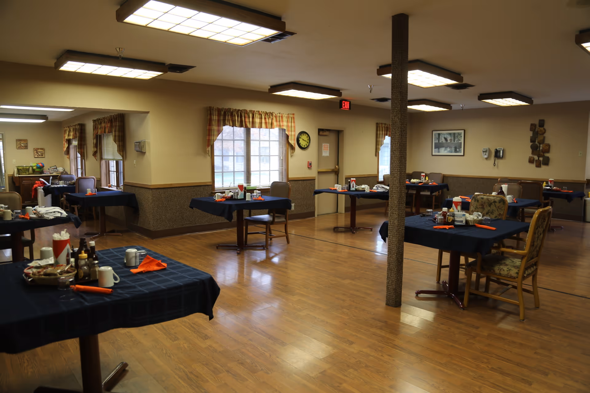 Spacious dining room with multiple tables set with dark tablecloths and place settings in a senior living facility.