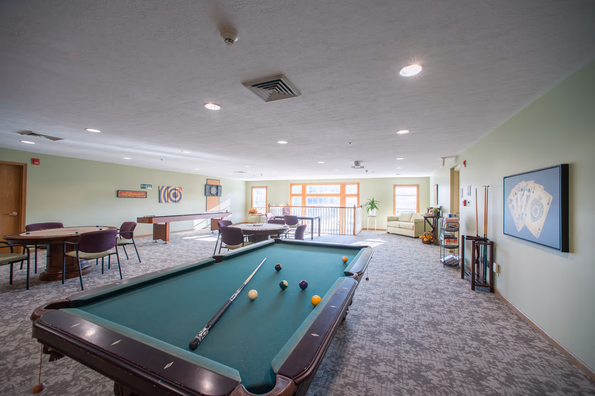 A spacious recreational room with a pool table in the foreground, a shuffleboard table and dartboard on the far wall, several round tables with chairs, a beige couch near large windows, and light green walls decorated with framed artwork and a rack holding pool cues.