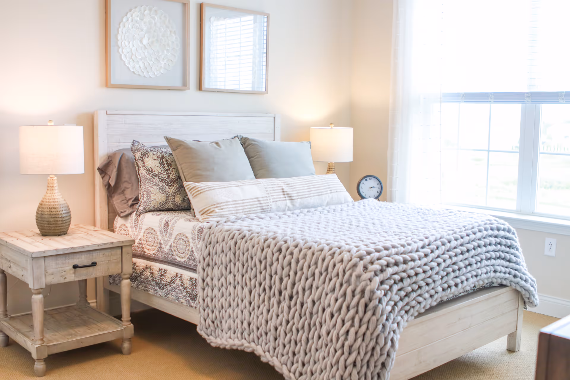 A bright bedroom with a wooden bed frame and a thick knitted gray blanket. The bed has multiple pillows with patterned and solid pillowcases. There are two matching wooden nightstands on either side of the bed, each with a lamp. A round clock is on the right nightstand. Two framed artworks hang on the wall above the bed. A large window with sheer curtains lets in natural light.