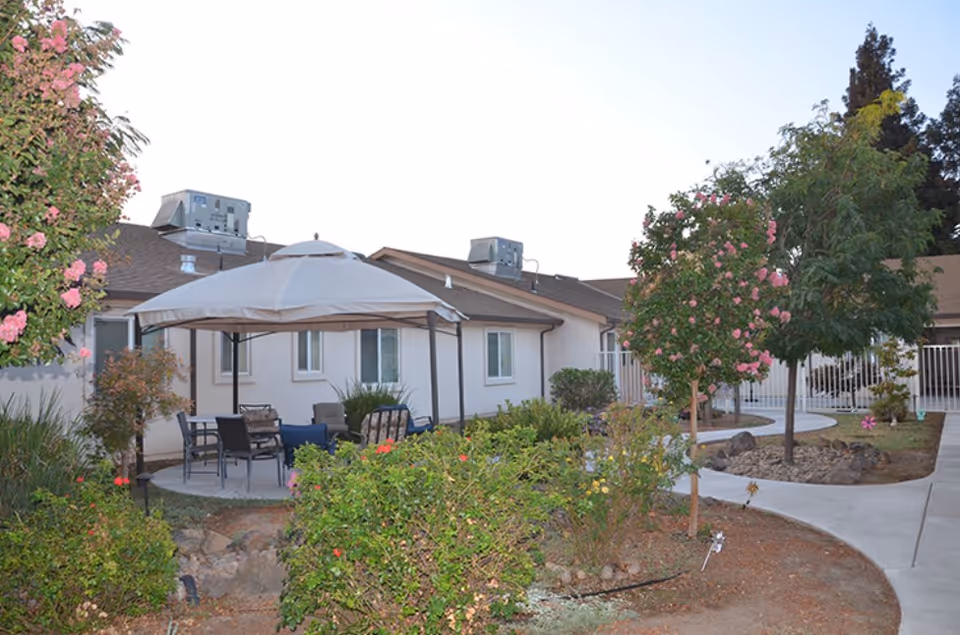Outdoor patio area at Hospitality House Assisted Living & Memory Care featuring a gazebo with chairs and a table surrounded by landscaped garden beds with flowering bushes and trees, adjacent to a single-story building.
