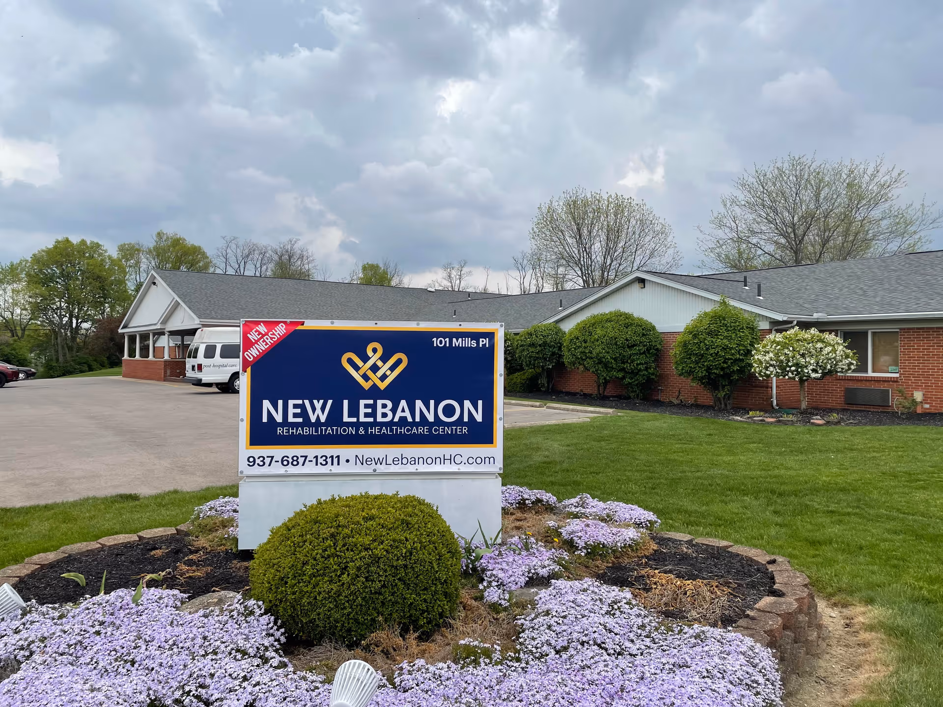 Entrance sign reading "New Lebanon Rehabilitation & Healthcare Center" in front of a single-story brick building with lawn and flowering groundcover.