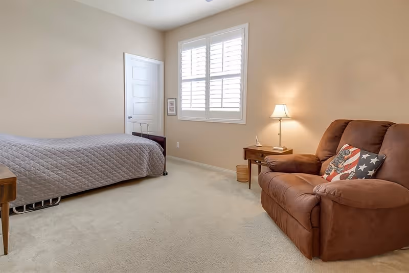 A simple bedroom with beige walls and carpet. There is a bed with a gray quilted bedspread on the left side, a window with white shutters in the center, and a brown recliner chair with a patriotic pillow on the right. A wooden side table with a lamp and a small wastebasket are next to the chair.
