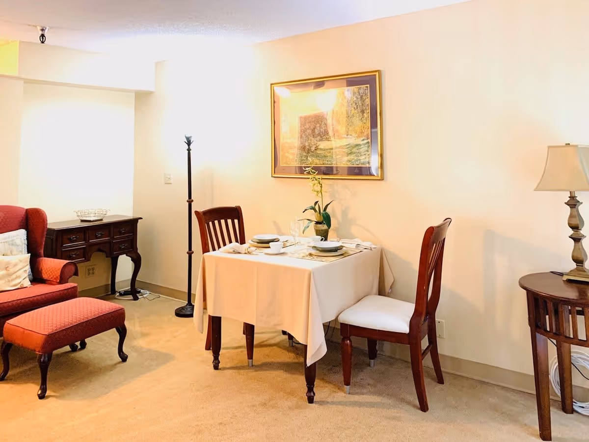 A cozy interior space featuring a small dining table set for two with white tablecloth, plates, cups, and a small plant centerpiece. To the left, there is a red upholstered armchair with a matching ottoman and a wooden side table with drawers. A tall floor lamp stands behind the dining table, and a framed painting hangs on the wall above it. On the right side, there is a wooden side table with a decorative lamp.