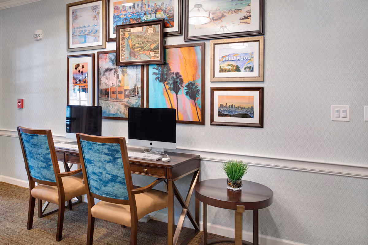 A small computer workstation area with two iMac computers on a wooden desk accompanied by two wooden chairs with blue upholstered backs and beige seats. The wall behind the desk is decorated with multiple framed pictures and artwork, including images of palm trees, a Hollywood sign, and cityscapes. A small round wooden side table with a green plant in a glass container is placed next to the desk. The room has light-colored patterned wallpaper and carpeted flooring.