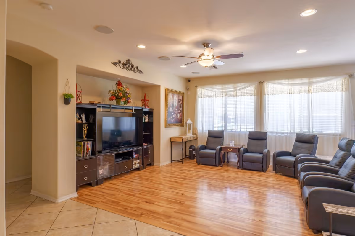 Bright communal living room with an entertainment center and TV, several reclining chairs, wood floor and large windows with sheer curtains.