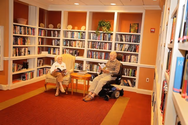 Two elderly individuals reading books in a cozy library room with bookshelves filled with books lining the walls. One person is seated in a striped armchair, and the other is in a motorized wheelchair. A small wooden table with a closed book is between them.