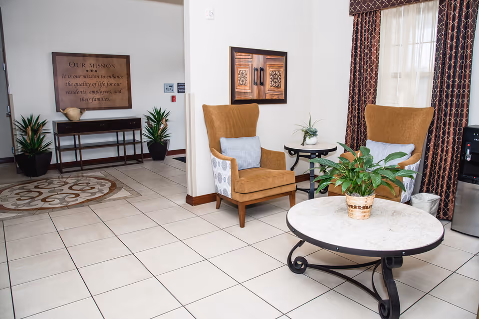 A cozy seating area in a senior living facility with two brown armchairs, each with a light blue cushion, positioned around a round marble-top coffee table with a potted green plant. Behind the chairs is a window with brown patterned curtains. To the left, there is a small table with decorative items and two potted plants on the floor. A framed mission statement is mounted on the wall above the table. The floor is tiled with a decorative circular pattern near the entrance.