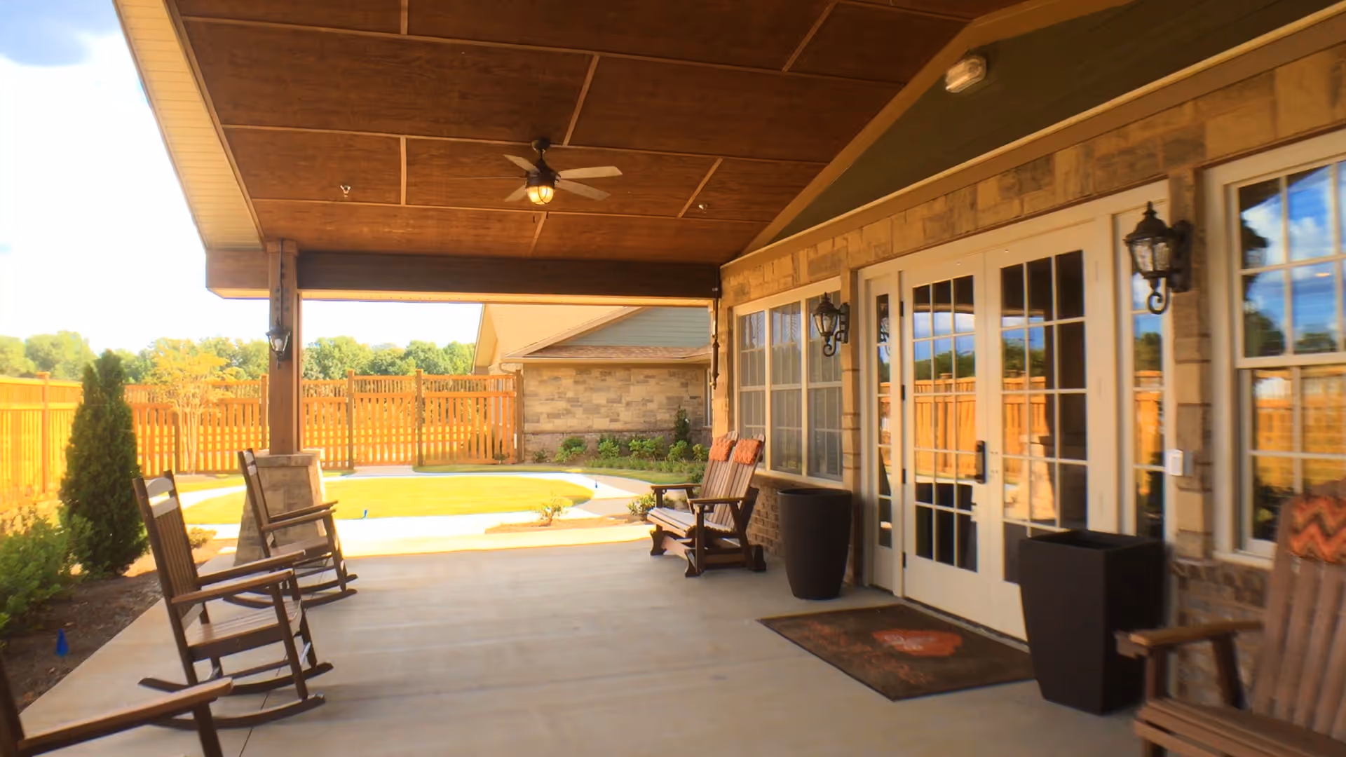 Covered outdoor patio area with wooden rocking chairs, ceiling fan, and glass double doors leading inside. The patio overlooks a fenced backyard with green grass and some shrubs.