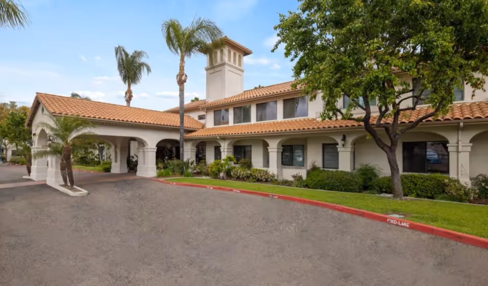 Exterior view of a senior living facility building with a tiled roof, arched entryway, palm trees, and landscaped greenery under a partly cloudy sky.
