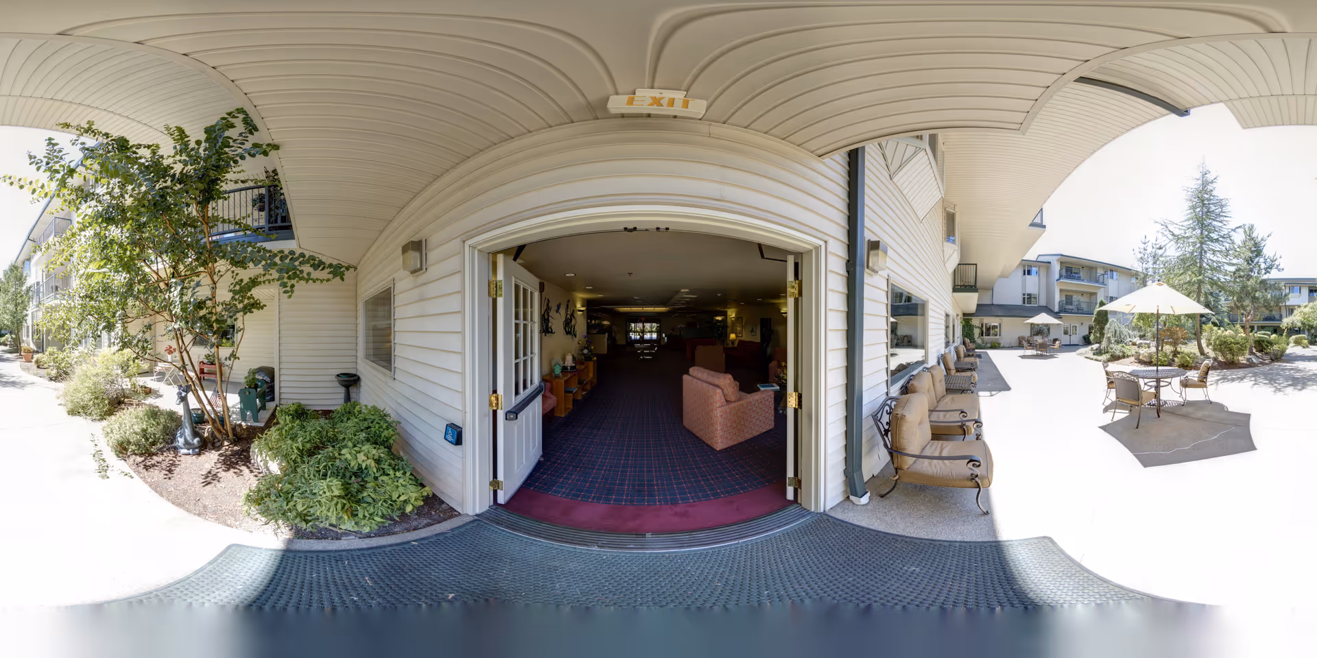 Open double doors leading from a lobby out to a sunny courtyard with patio tables, umbrellas, and building balconies.