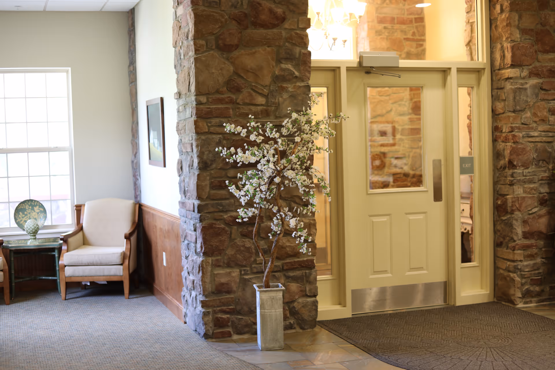Interior view of a common area with stone pillars, a glass door with an exit sign, a potted flowering plant, and two beige armchairs next to a window with a decorative plate on a small table.