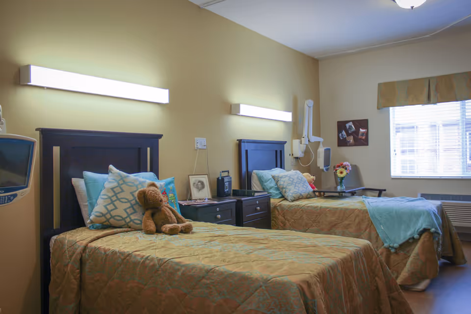 Two neatly made twin beds in a well-lit senior facility bedroom with bedside tables, medical equipment, and a teddy bear on one bed.