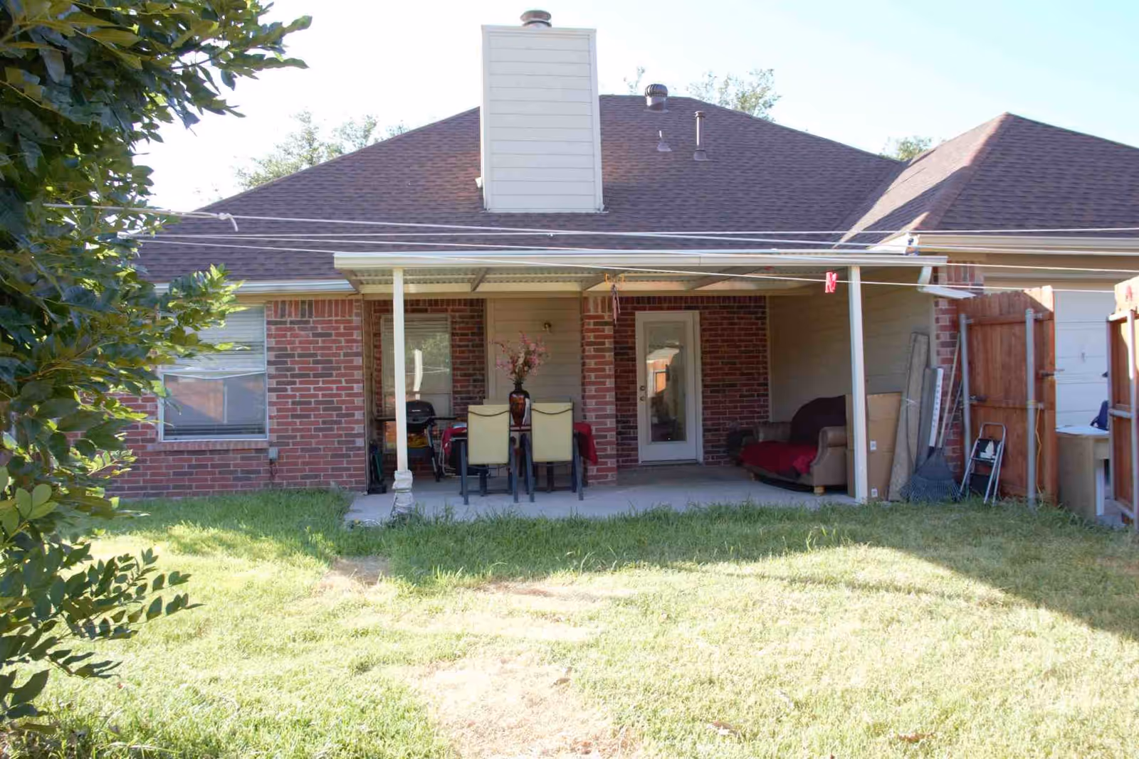 Backyard view of a single-story brick house with a covered patio area. The patio has a small dining table with four chairs and a vase with flowers on top. There is a couch with a red blanket on the right side of the patio. The yard has green grass and some bushes on the left side. A wooden fence and a garage door are visible on the right side.