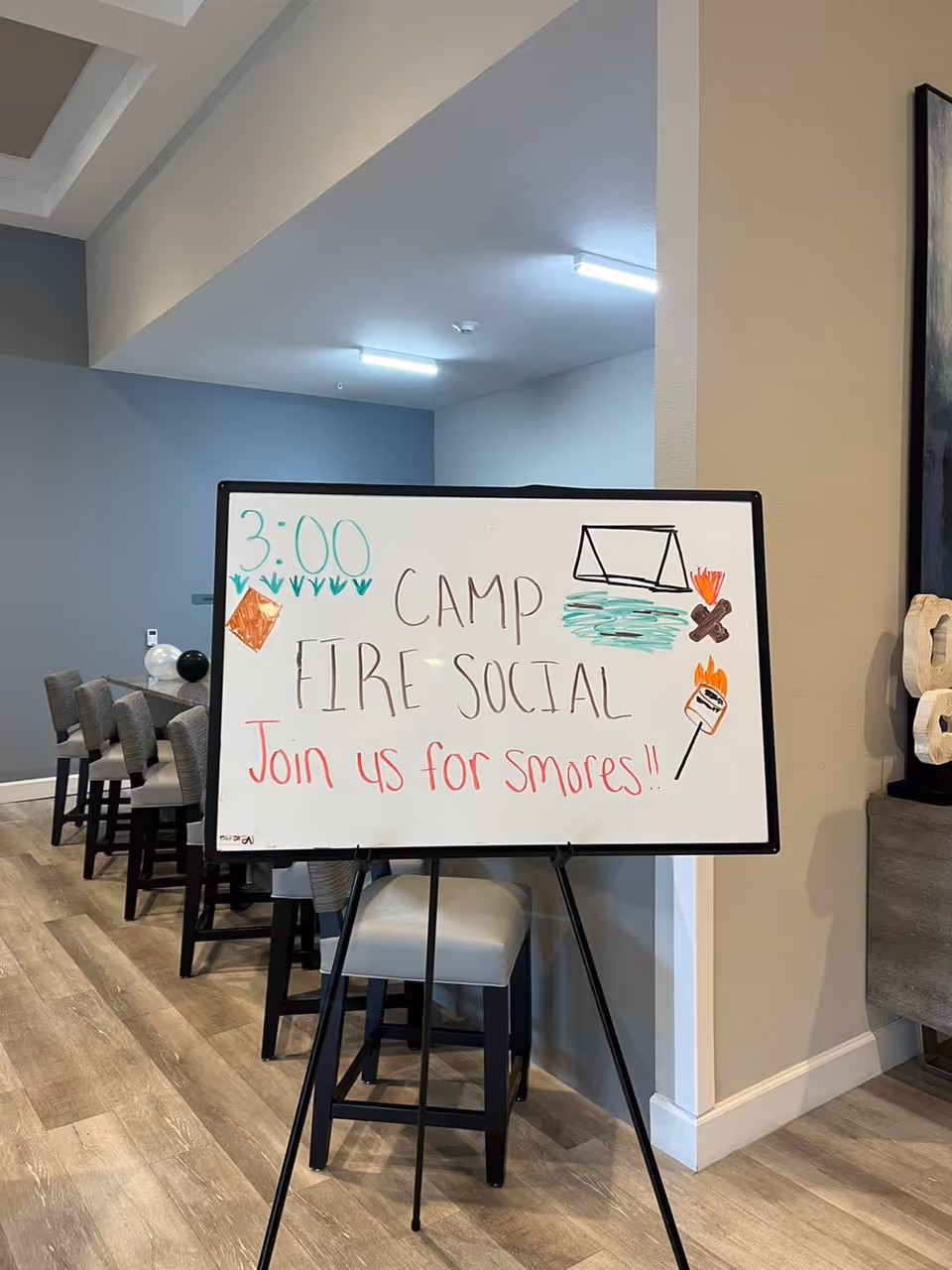 A whiteboard easel in a senior living common area announcing a 'Camp Fire Social' and s'mores, with high stools and a dining counter behind it.