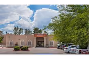 Exterior view of a single-story building with a sign reading 'Ahwatukee Post Acute' above the entrance. The building is surrounded by trees and bushes, with several parked cars visible on the right side under a partly cloudy sky.