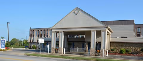 Exterior front view of Cardinal Nursing and Rehabilitation facility showing a covered entrance with columns, a sign on the left side, and a clear blue sky.
