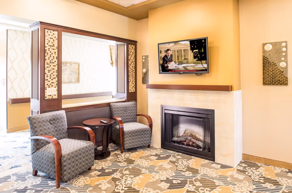 A cozy seating area with two patterned armchairs and a small round wooden table between them, placed on a floral-patterned carpet. A modern electric fireplace is set into a beige wall, above which a flat-screen TV is mounted showing a man in a suit reading papers. The room has warm yellow walls and decorative wall art on either side of the fireplace. A large window opening with wooden trim looks into an adjacent room with similar wallpaper and a framed picture.
