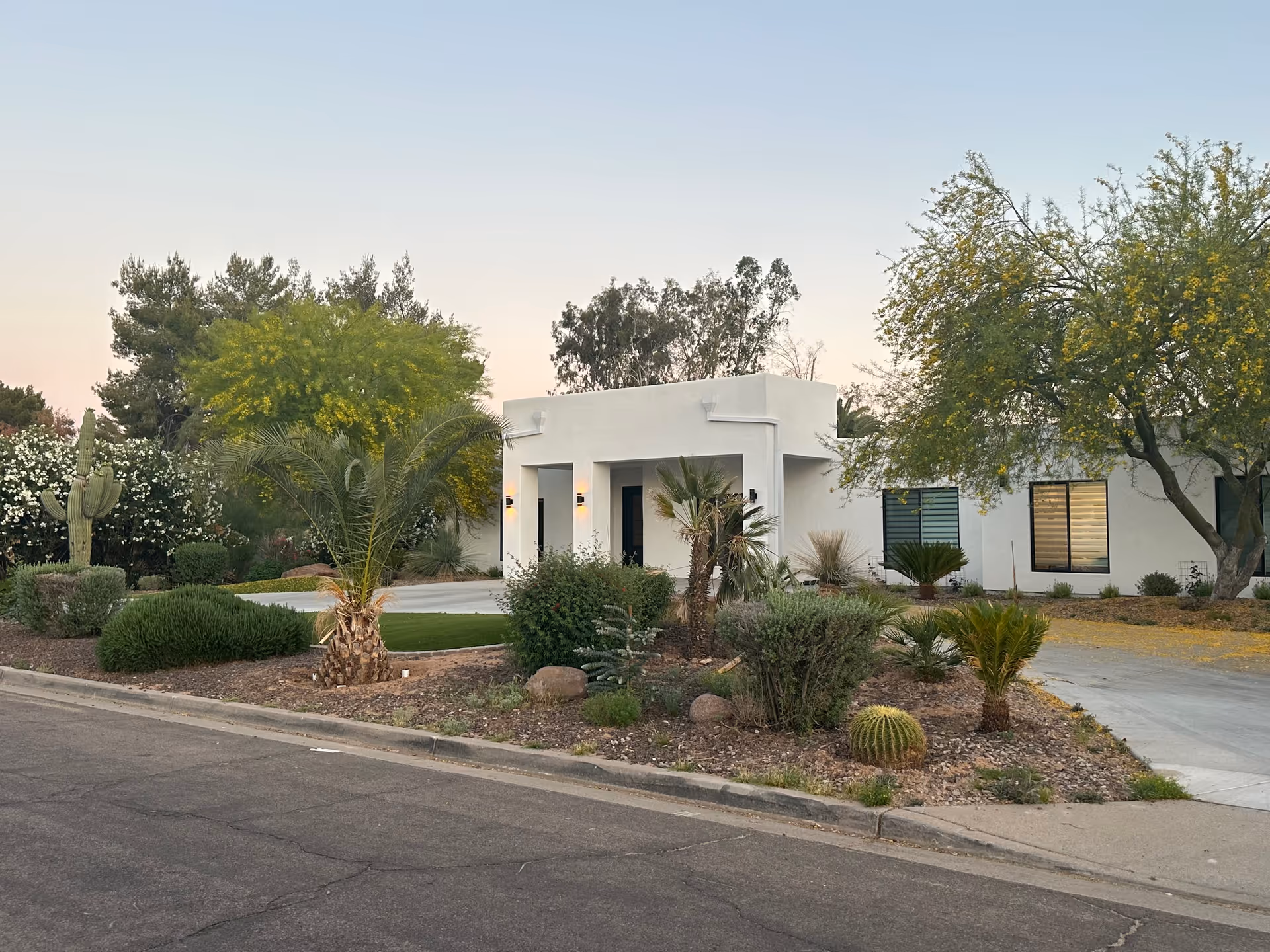 Exterior view of OakBrook Assisted Living facility showing a single-story white building with a flat roof, surrounded by desert landscaping including palm trees, bushes, cacti, and other plants, with a paved driveway and street in the foreground under a clear sky.