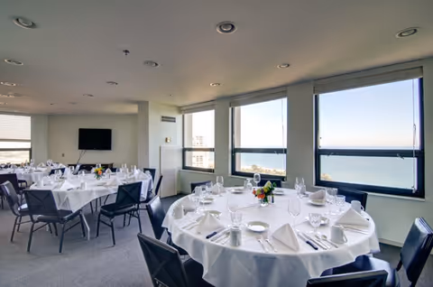 A bright dining room with round tables covered in white tablecloths, set with glassware, plates, napkins, and silverware. The room has large windows offering a view of the ocean and blue sky. Several black chairs surround each table, and a flat-screen TV is mounted on the far wall.