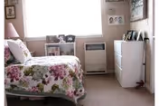 A small bedroom in a senior residence with floral bedspread, dresser, and window.