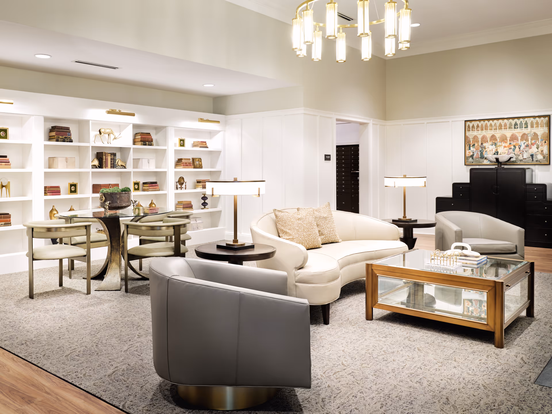 A bright and elegant senior living common area featuring a curved white sofa with two decorative pillows, two gray armchairs, a glass coffee table with a wooden frame, and two side tables with lamps. In the background, there is a white built-in bookshelf filled with books and decorative items, a round table with four chairs, and a large framed artwork on the wall. The room has a patterned carpet and a modern chandelier hanging from the ceiling.