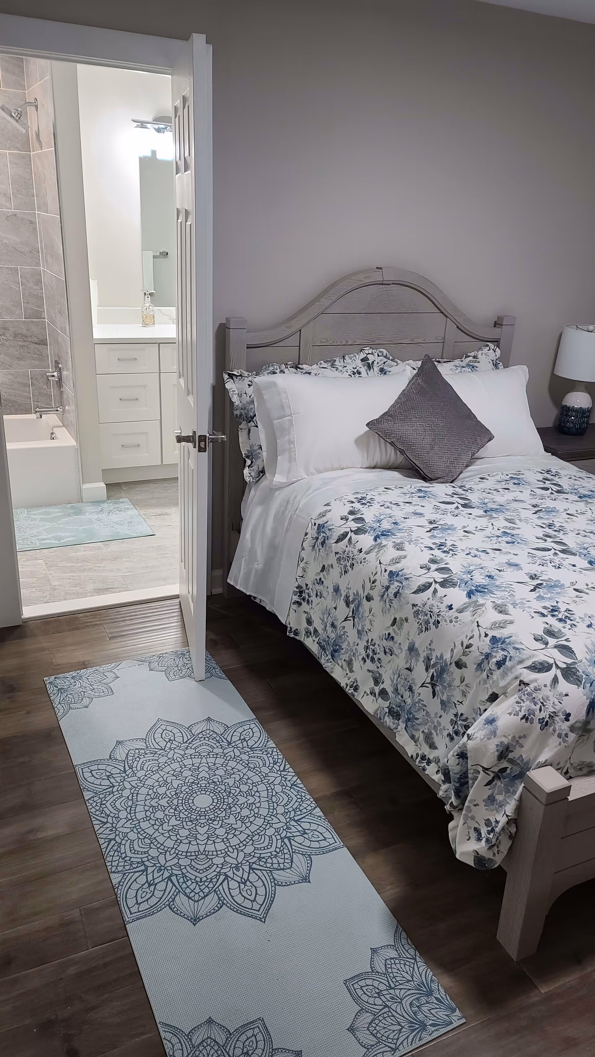 A bedroom with a wooden bed frame, floral bedding, and a gray decorative pillow. A door is open showing an adjacent bathroom with a bathtub, tiled walls, a white vanity with drawers, and a mirror above the sink. A patterned rug is placed on the wooden floor leading from the bedroom to the bathroom.