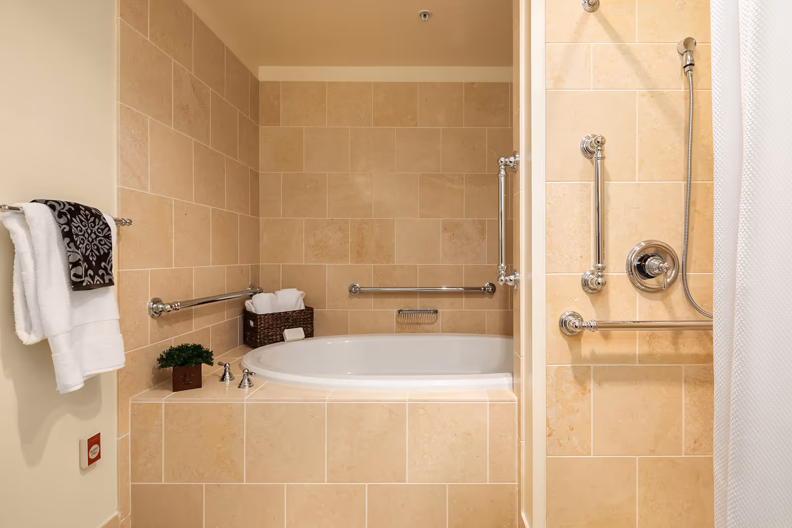 A bathroom with beige tiled walls and a built-in bathtub. The bathtub area has multiple chrome grab bars for safety. There is a basket with rolled white towels on the edge of the tub and a small potted plant. A towel rack on the left wall holds two white towels and a decorative black and white towel. A shower area with a chrome showerhead and grab bars is partially visible on the right side.