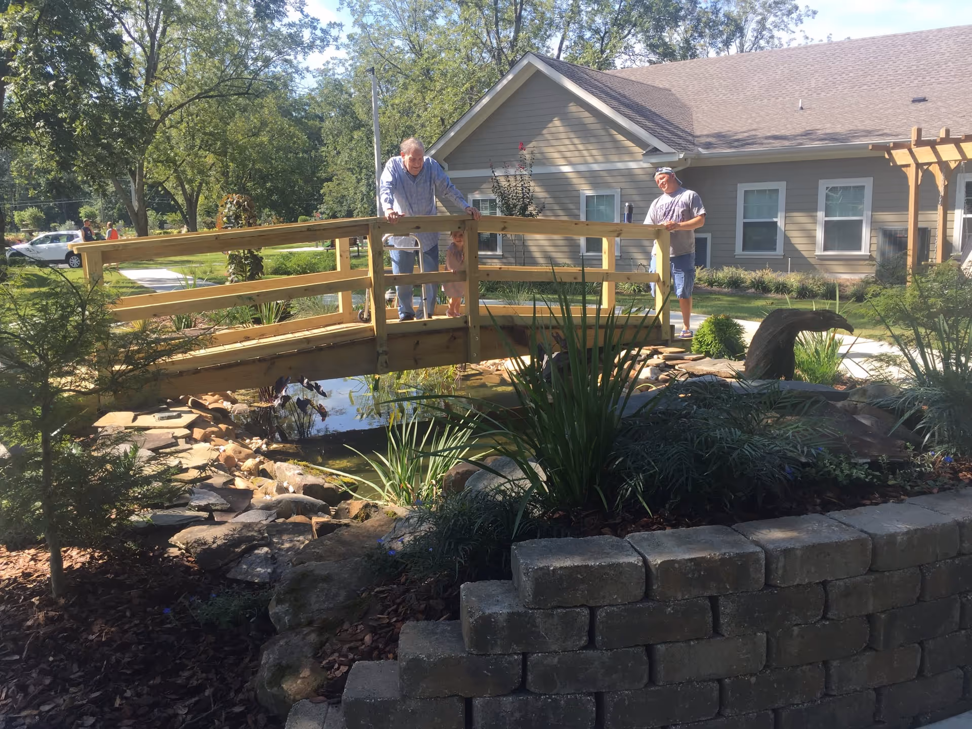 An elderly man with a walker and a young child standing on a wooden bridge over a small pond in a garden area. A man in casual clothes stands nearby on the path. Behind them is a beige building with white-trimmed windows and a pergola. The garden includes various plants, rocks, and a small water feature.